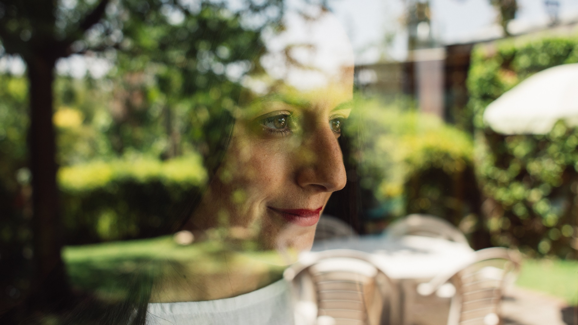 Vrouw met donker haar en wit shirt kijkt door het raam naar de tuin