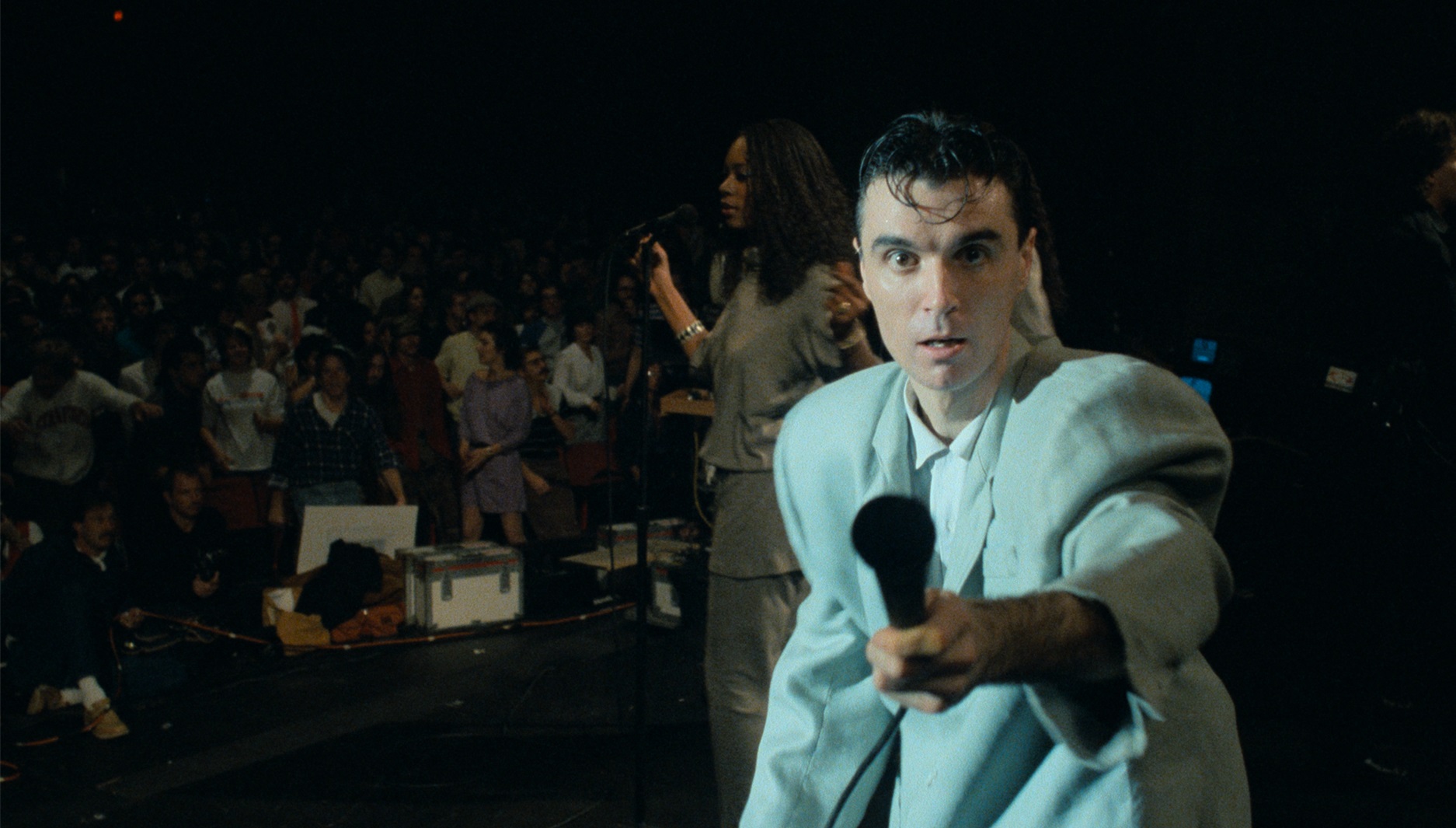 Zanger David Byrne heeft een wit pak aan en wijst met zijn microfoon naar de fotograaf